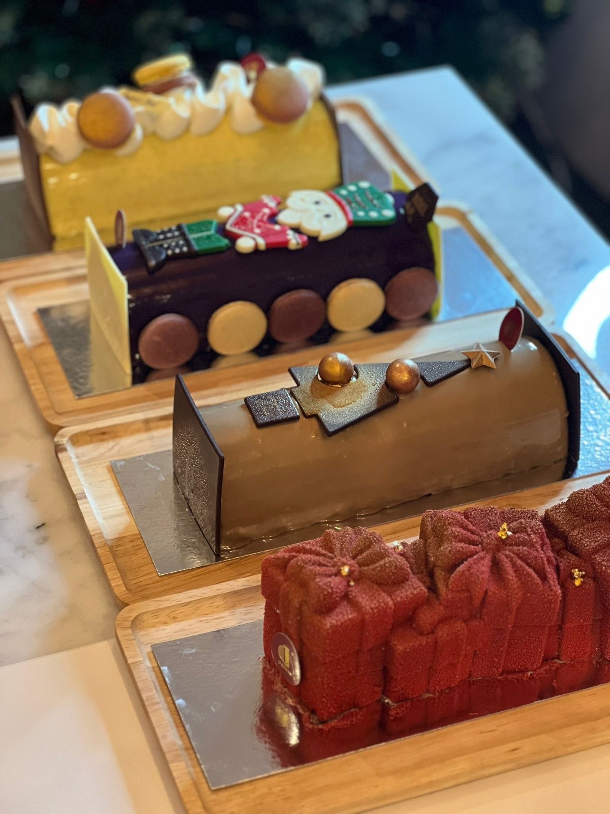 Quatre bûches de Noël signature : agrumes et macaron, chocolat noir au décor bonhomme de neige, praliné au sapin doré, et bûche en trompe-l'œil paquets cadeaux velours rouge